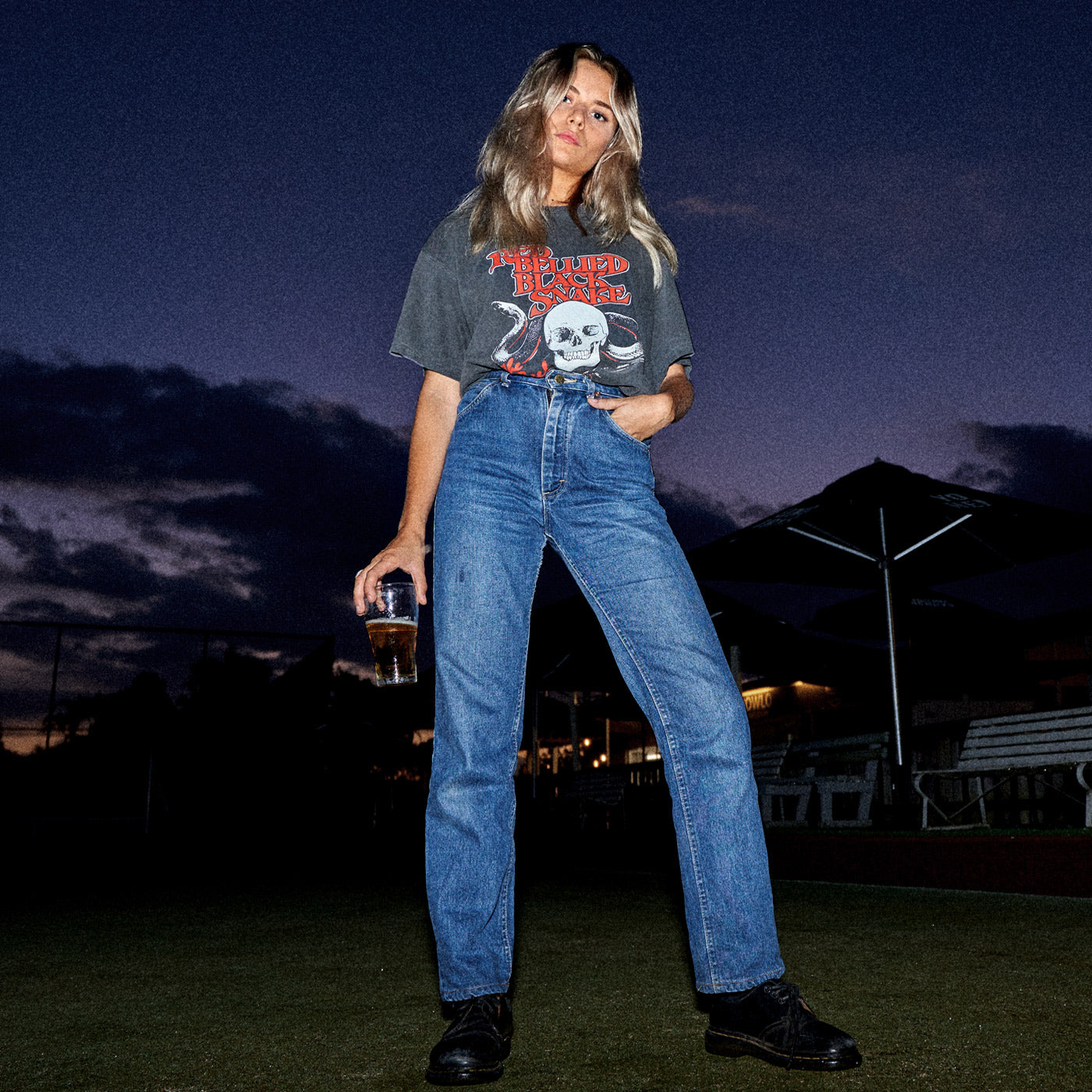 A lady standing on a lawn bowls ground holding a drink in The Beefs Red Bellied Black Snake t-shirt