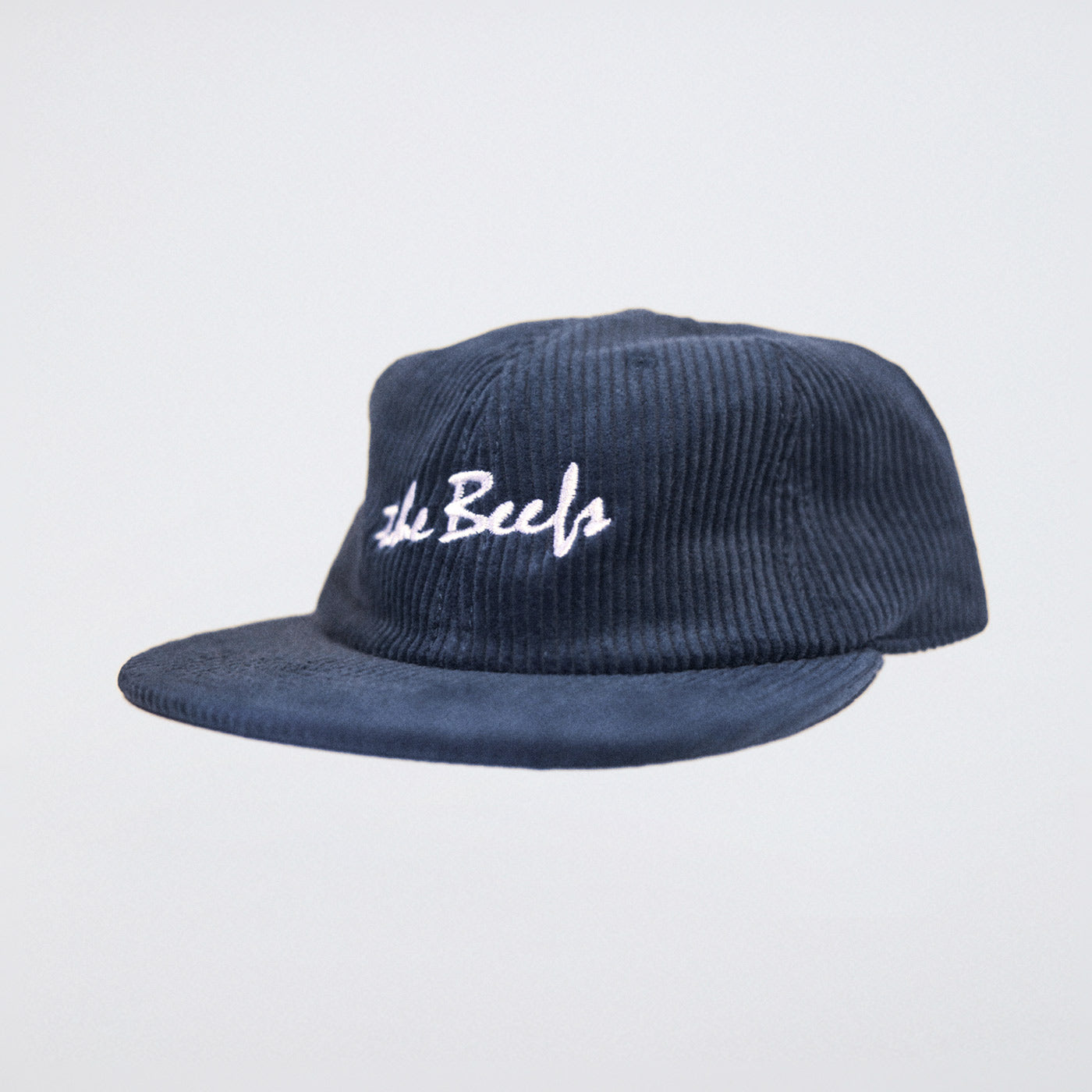 A steel blue cord hat with The Beefs logo photographed in a studio on an angle