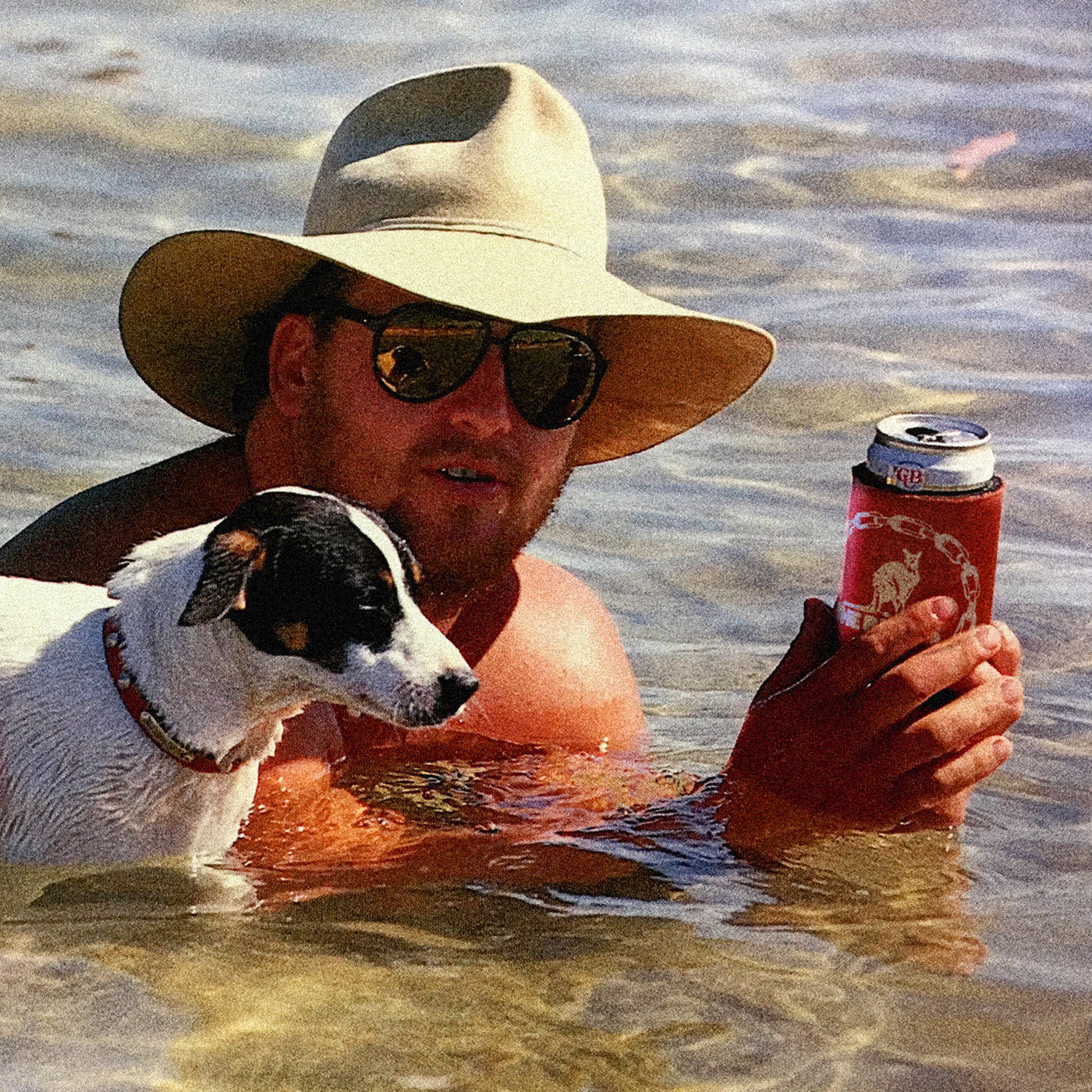 An Australian man in an Akubra swimming at the beach with his dog